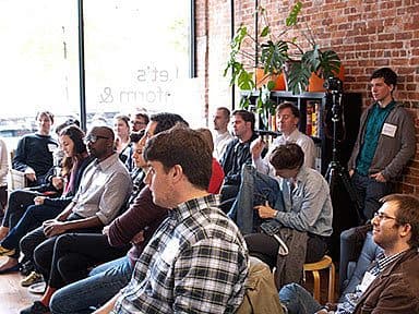 A group of people seated and standing in a casual indoor setting with brick walls and large windows. Some are taking notes, while others are listening attentively. Potted plants and a shelf with items are visible. The atmosphere appears to be relaxed and engaging.