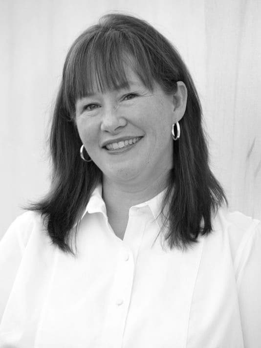 Black and white portrait of a smiling woman with shoulder-length dark hair and bangs. She is wearing a light-colored button-down shirt and hoop earrings. The background is neutral.