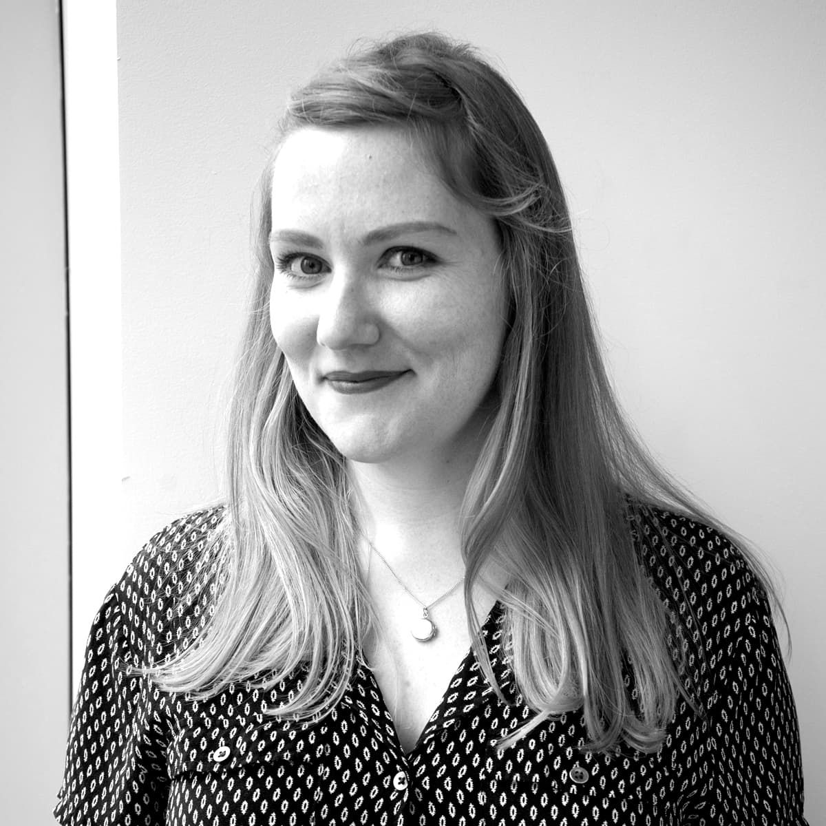 Black and white image of a woman with long, straight hair, smiling slightly, and facing the camera. She is wearing a patterned blouse and a necklace with a pendant. The background is a plain wall.