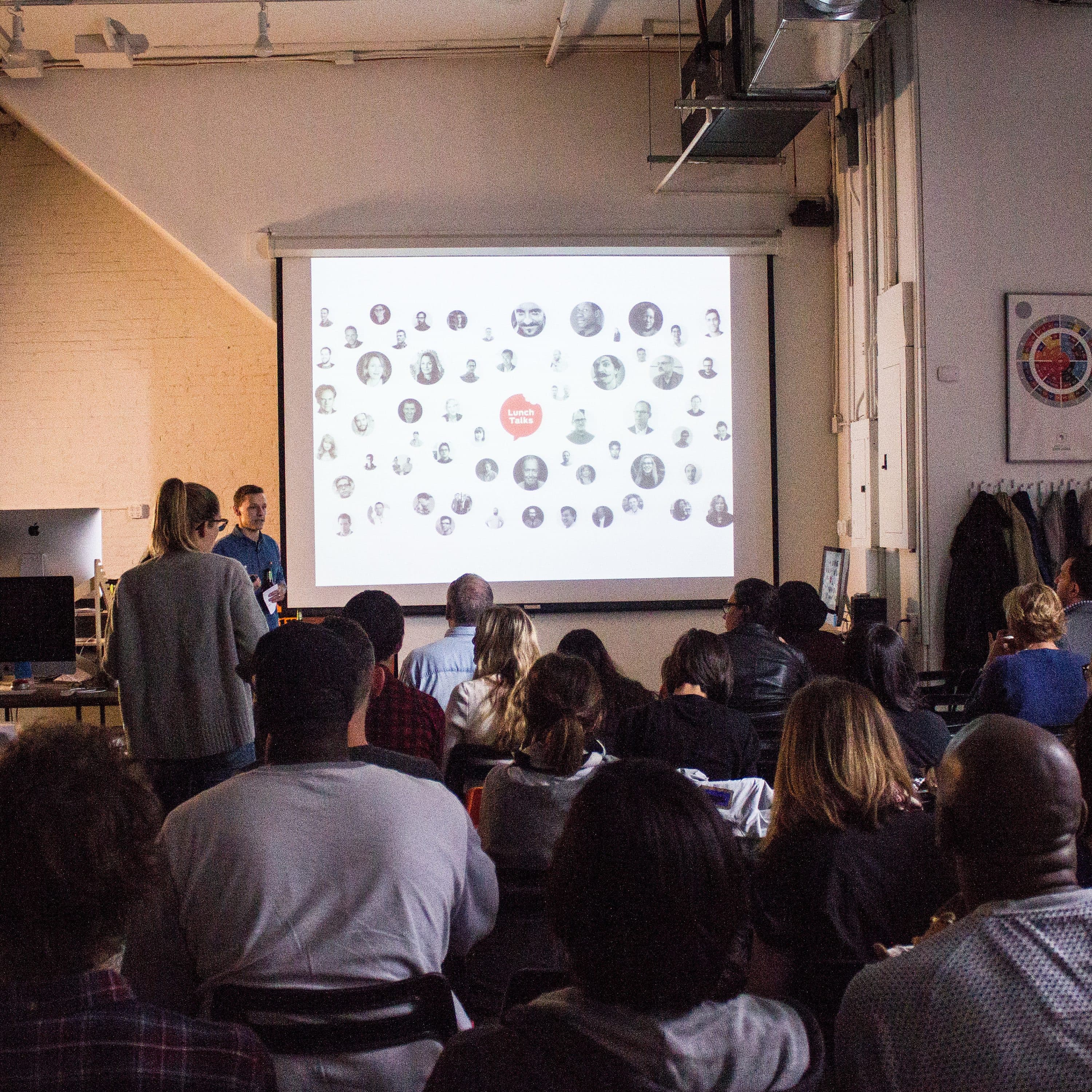 A group of people are seated in a dimly lit room, attentively watching a presentation. The presenter is standing near a screen displaying a collage of various portraits and icons. Shelves, a coat rack, and wall art are visible in the background.