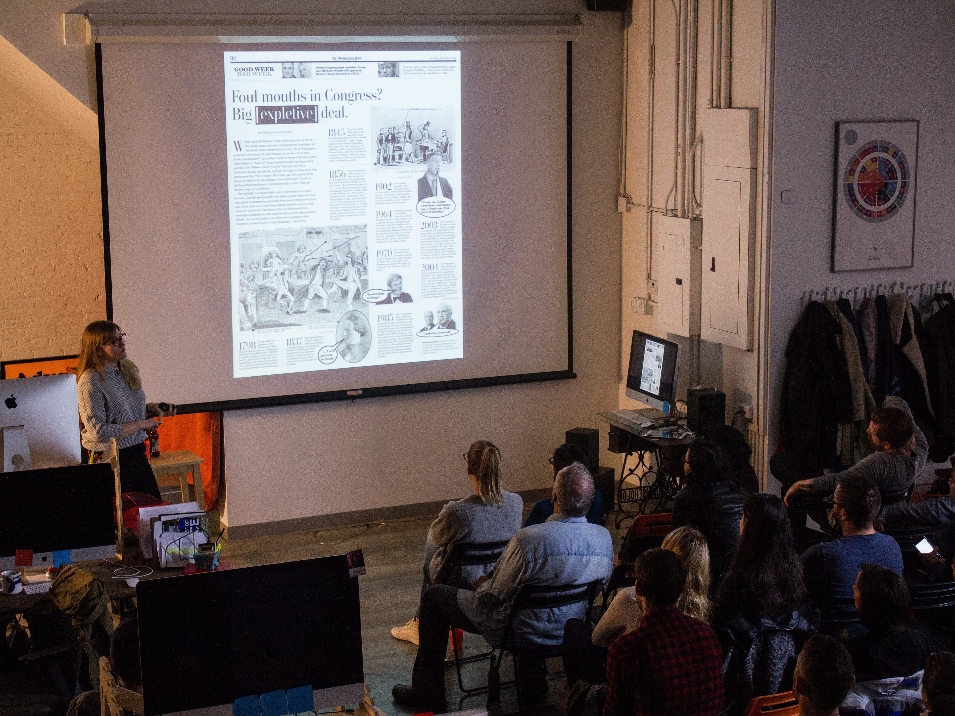 A person stands at the front of a room, giving a presentation to a group of seated attendees. A large projection screen displays a detailed document or webpage. The room has a modern, industrial design with exposed brick, a color wheel on the wall, and various computer equipment.
