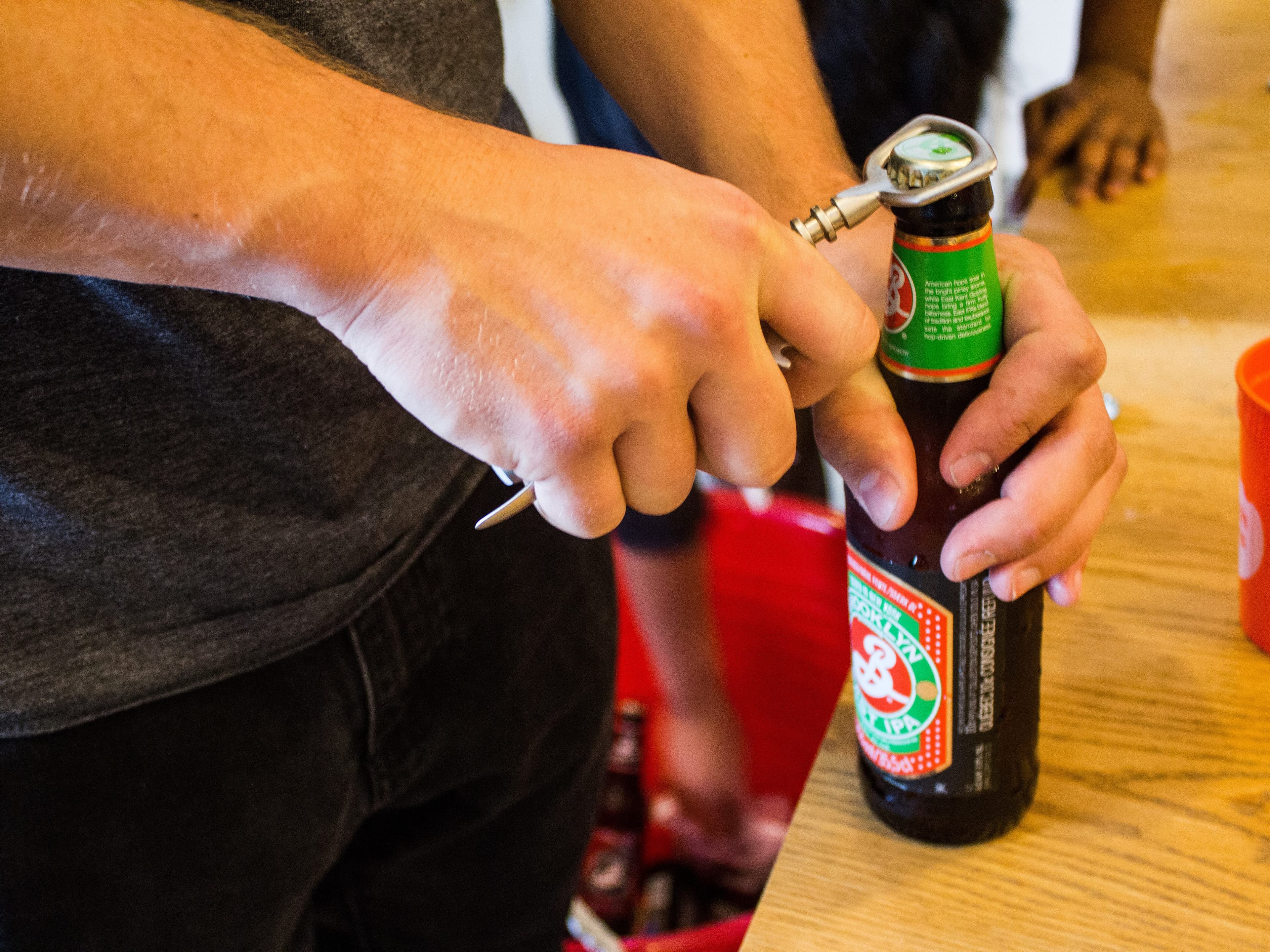 A person in a grey t-shirt uses a bottle opener to open a brown glass beer bottle on a wooden table. In the background, another person reaches into a red bucket filled with more beer bottles. An orange cup is also visible on the table.