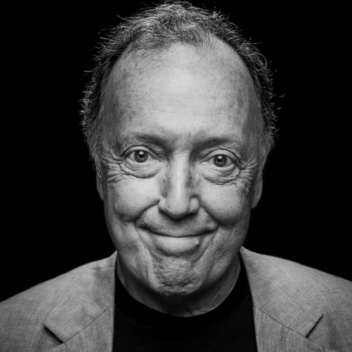 A grayscale portrait of an older man with thinning hair, giving a pleased smile. He is dressed in a light-colored blazer over a dark shirt. The background is solid black, focusing attention on his face and expression.