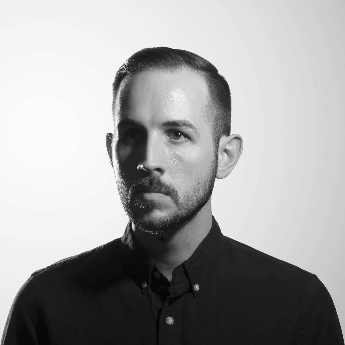 Black and white portrait of a man with short, neatly combed hair and a full beard. He is wearing a buttoned-up shirt and looking slightly to the side with a neutral expression. The background is plain and well-lit.