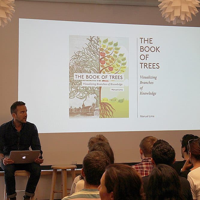 A man sits on a stool in front of a screen giving a presentation to an audience. The slides on the screen are titled "The Book of Trees: Visualizing Branches of Knowledge" by Manuel Lima, showcasing the book cover and details about the work.