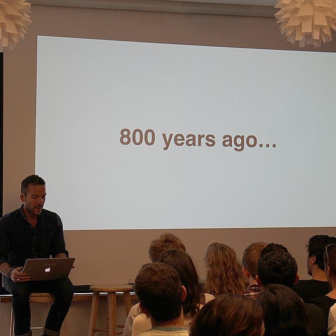 A man is sitting and presenting to an audience in a brick-walled room. The screen behind him displays the text "800 years ago...". He is holding a laptop, and the audience members are seated and listening attentively.