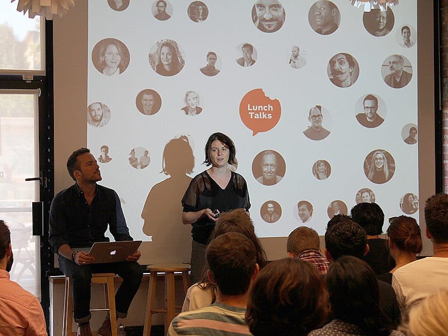 A woman and a man are presenting at a "Lunch Talks" event. The man sits with a laptop while the woman stands and speaks to an audience. Behind them, a projection screen displays a collage of various headshots inside circular frames.