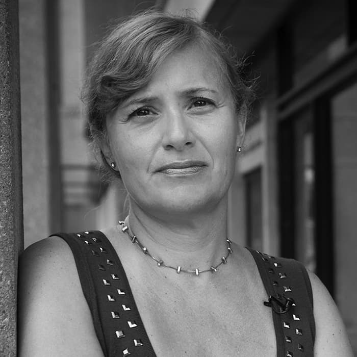 A woman with short, wavy hair stands against a column, wearing a dark sleeveless top adorned with small square studs. She has a thoughtful expression and is wearing a beaded necklace. The background shows an urban setting with buildings and shadows. The photo is in black and white.