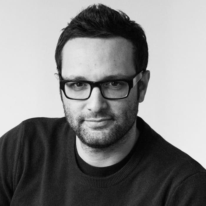 A man with short dark hair, wearing black-framed glasses and a dark sweater, looks directly at the camera with a slight smile. The background is plain and white, highlighting his face and attire.