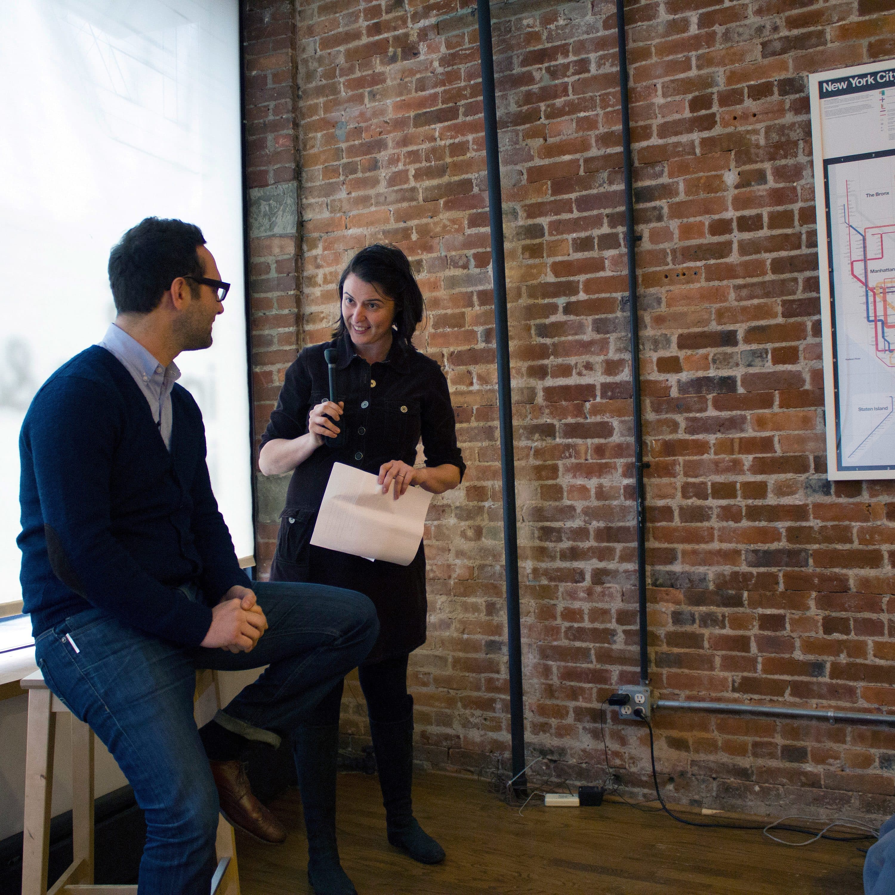 A person holding a microphone is speaking to another person seated on a stool by a window in a brick-walled room. There is a New York City Subway Diagram on the wall behind them. The speaker holds a piece of paper, and there is a sandwich on a plate in the foreground.