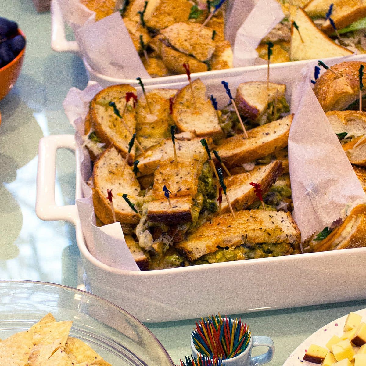 A table set for a party with platters of sandwiches held together with colorful toothpicks, bowls of purple and red grapes, a plate of tortilla chips, and cubes of cheese. The sandwiches are placed in white ceramic dishes lined with parchment paper.