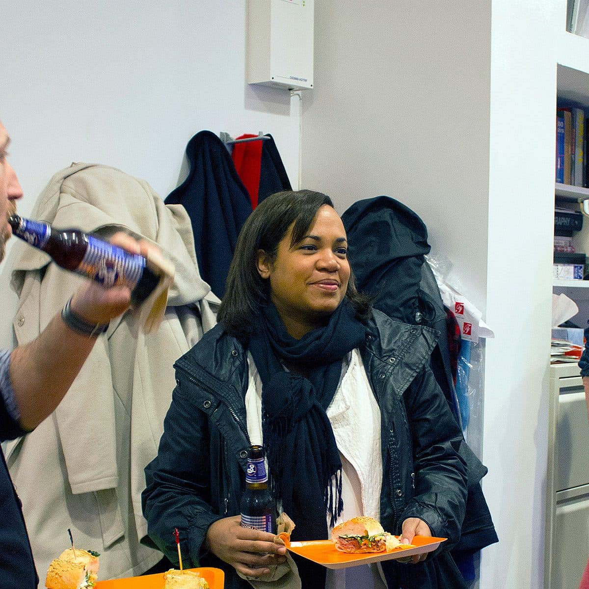 Three people are socializing in an indoor space, holding drinks and plates with sandwiches. A bald man in a black sweater is drinking from a bottle, a woman in a black jacket is smiling, and a man in glasses and a red shirt is conversing with them. Coats hang in the background.