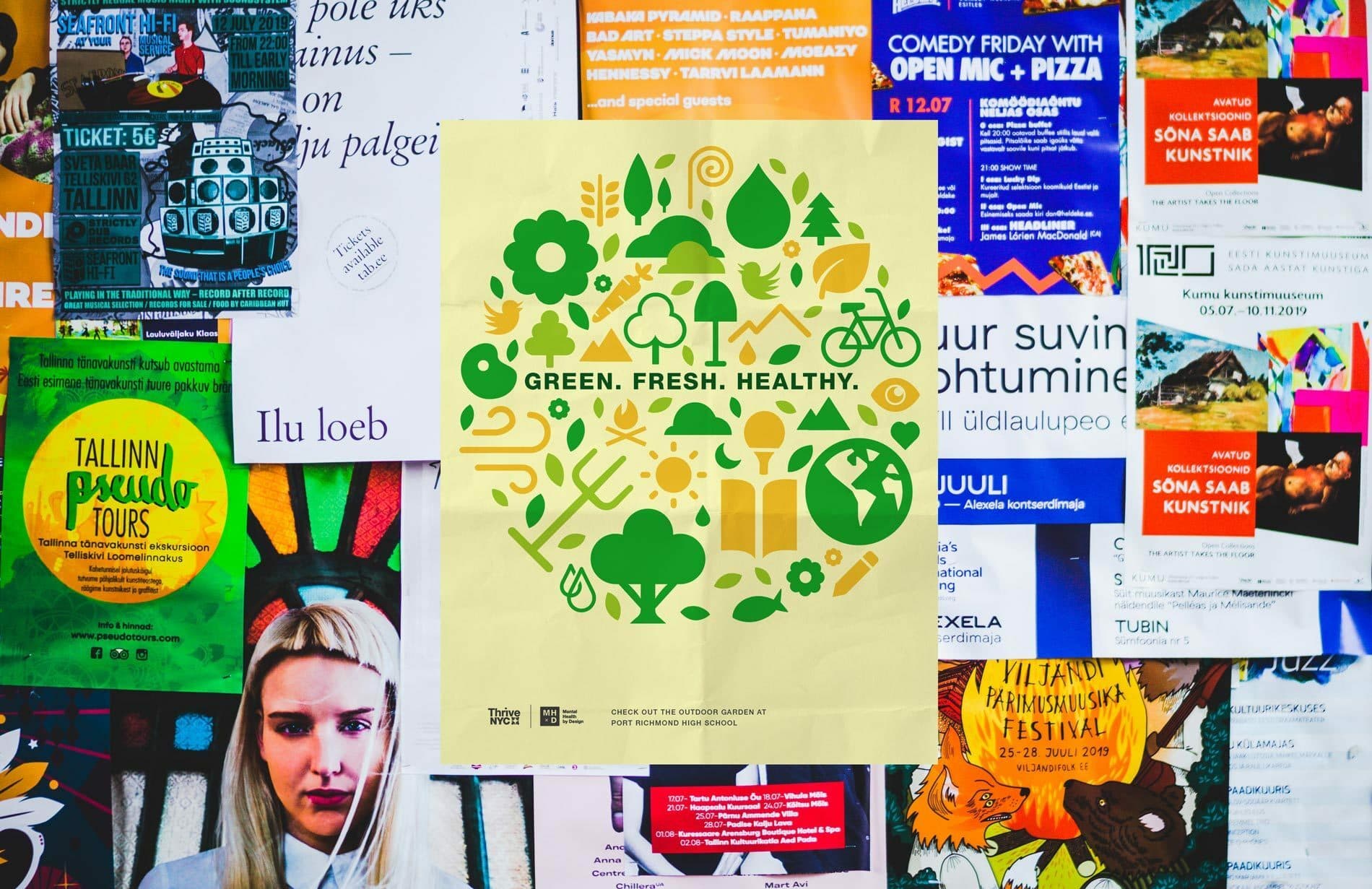 A collage of various colorful posters and flyers on a wall. In the center is a green-themed poster featuring icons of plants, trees, and bicycles with the text "GREEN. FRESH. HEALTHY." Surrounding it are posters advertising events, tours, and performances.