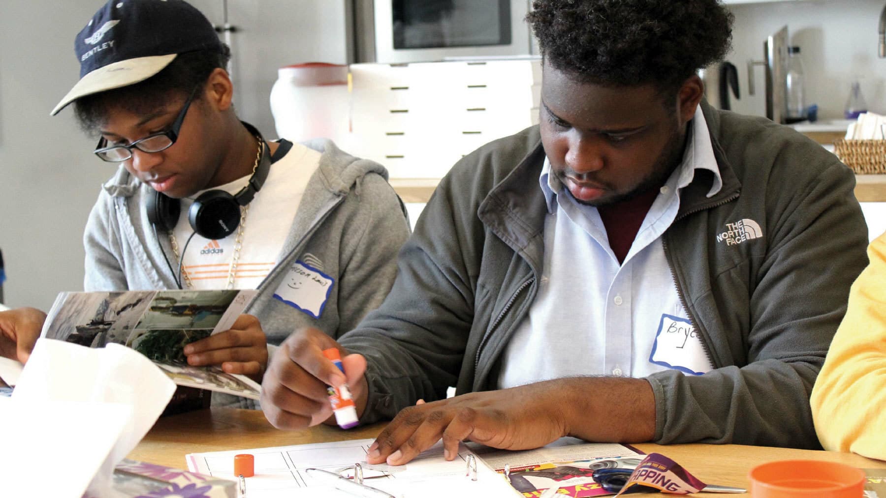 Two young men are seated at a table engaged in crafting activities. One is holding a magazine and the other is using a glue stick while focusing on a piece of paper. Both wear name tags and casual clothing. Various craft supplies are scattered on the table.