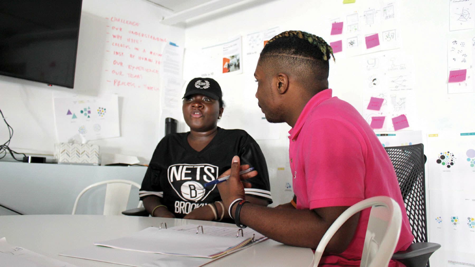 Two people sit at a table in a brightly lit office. One wears a Brooklyn Nets jersey and the other a pink shirt. They are talking, with papers and notebooks in front of them. Behind them, the wall is covered with various charts, sticky notes, and diagrams.