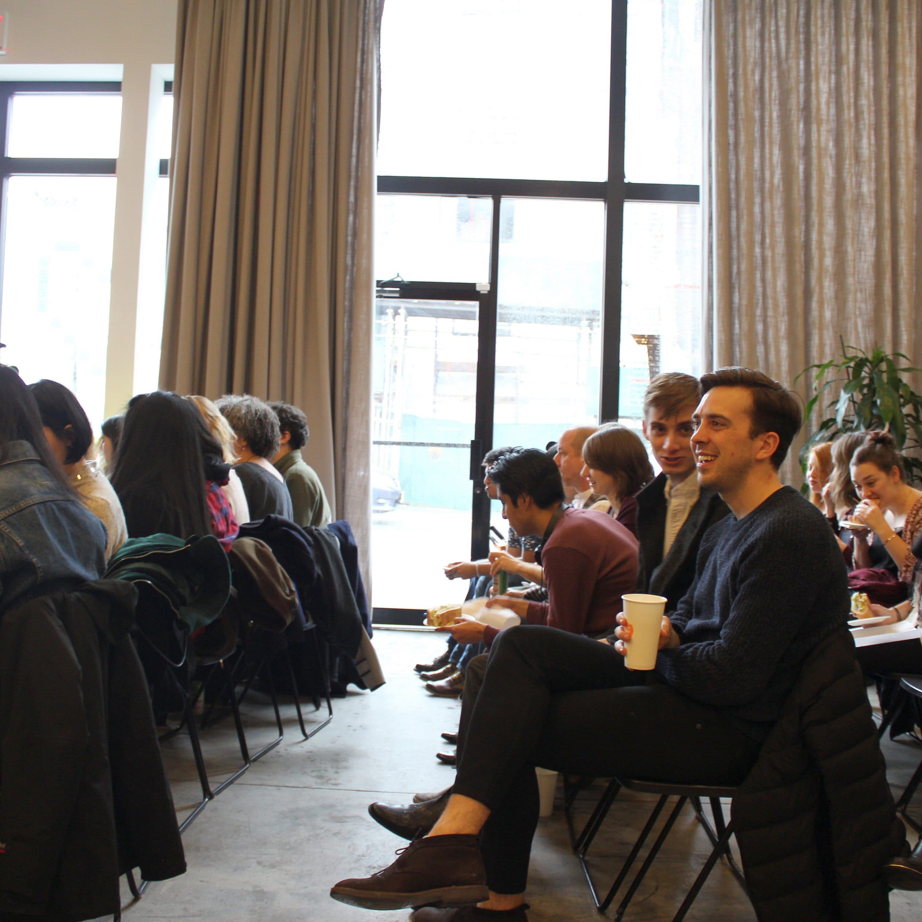 A group of people sitting in rows of chairs, facing the same direction, and engaging in conversation. Some are smiling and holding coffee cups. Large windows in the background let in natural light, and a few plants are visible near the windows.