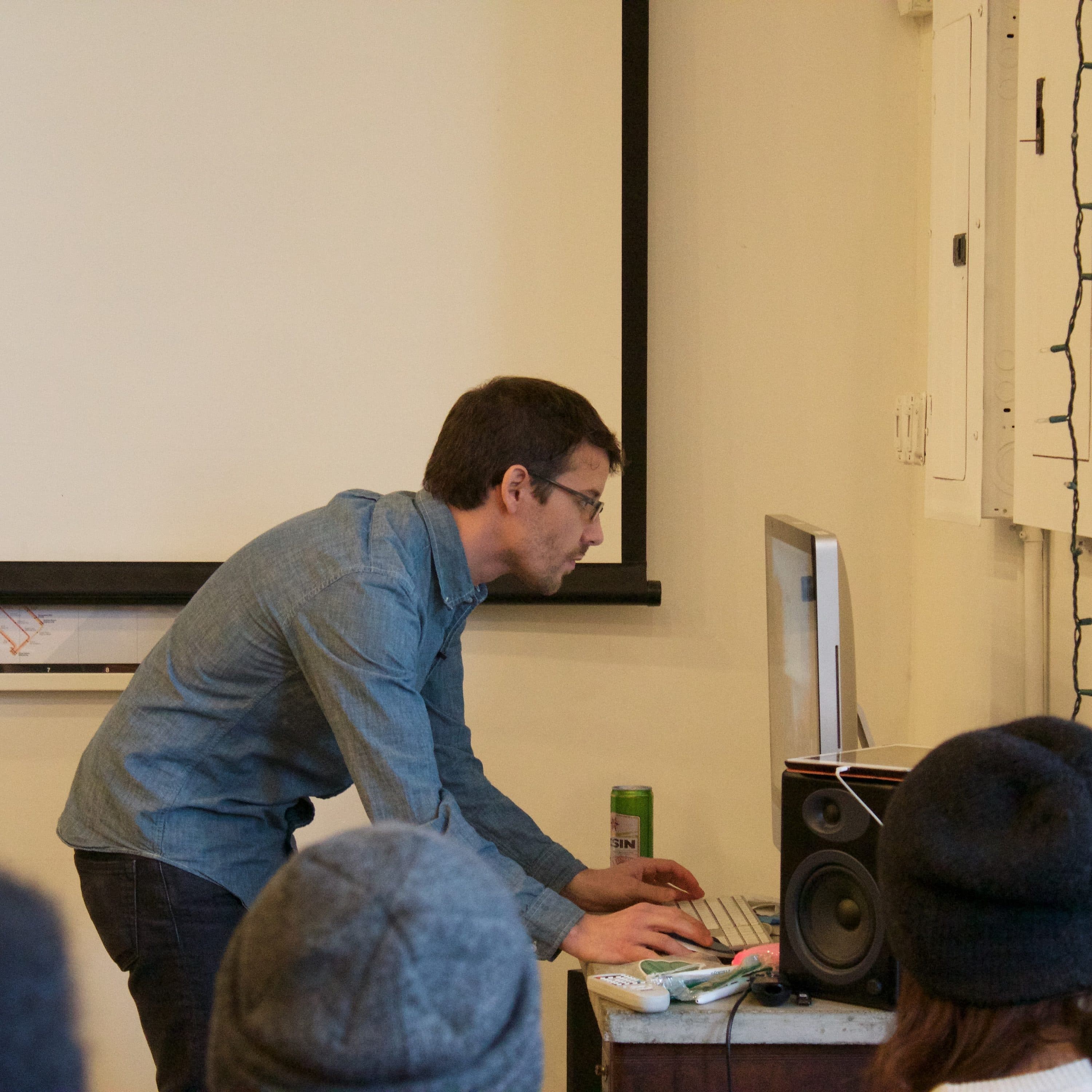 A person wearing glasses and a blue shirt is standing and typing on a computer keyboard at a desk. They are facing a monitor with a speaker next to it. Several people wearing hats are seated facing the person. A projector screen and string lights are visible.