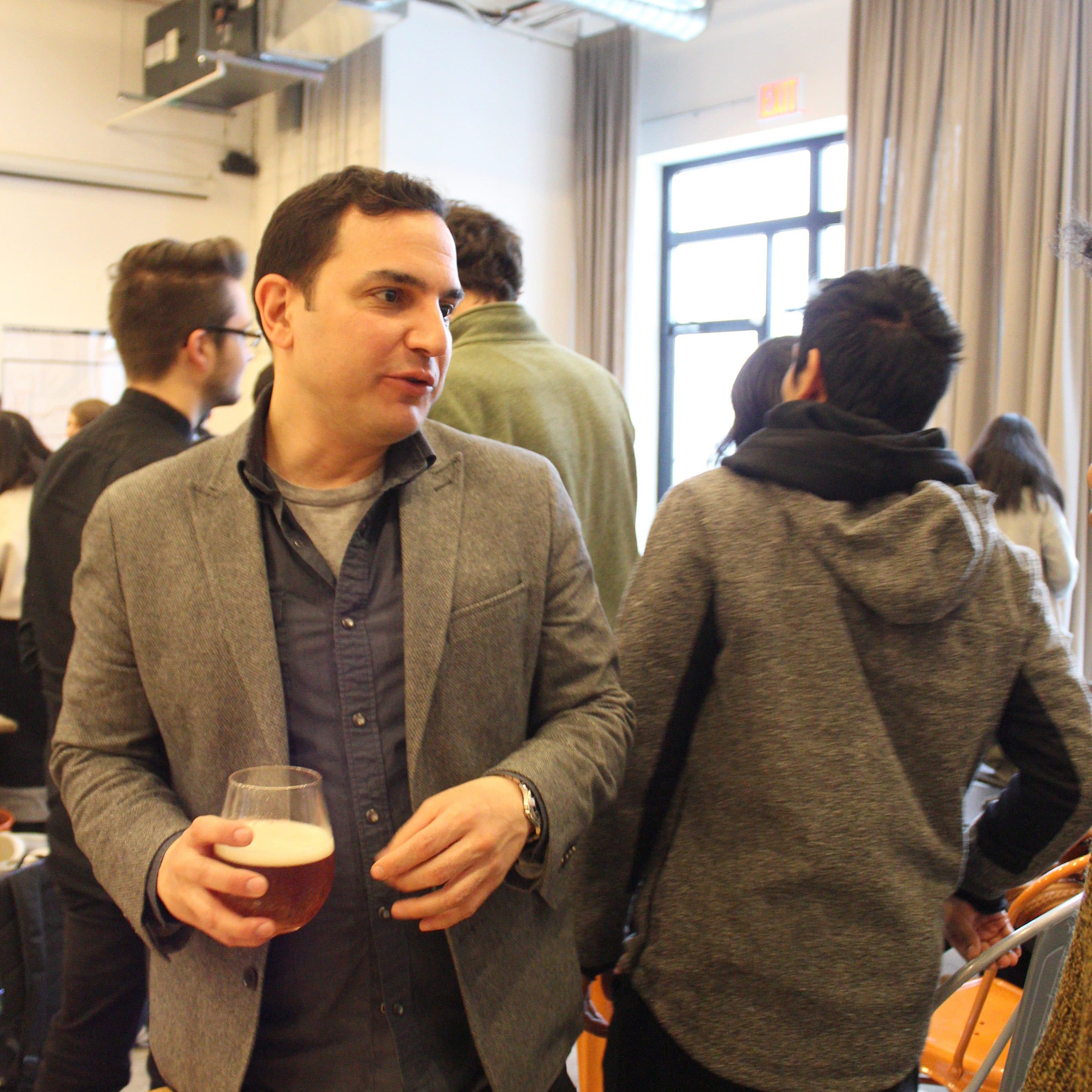 A man in a gray blazer and blue shirt holds a glass of beer while conversing at a social gathering. Behind him, several people are engaged in conversation in a room with large windows, curtains, and modern industrial decor. A can of beer is on the table nearby.