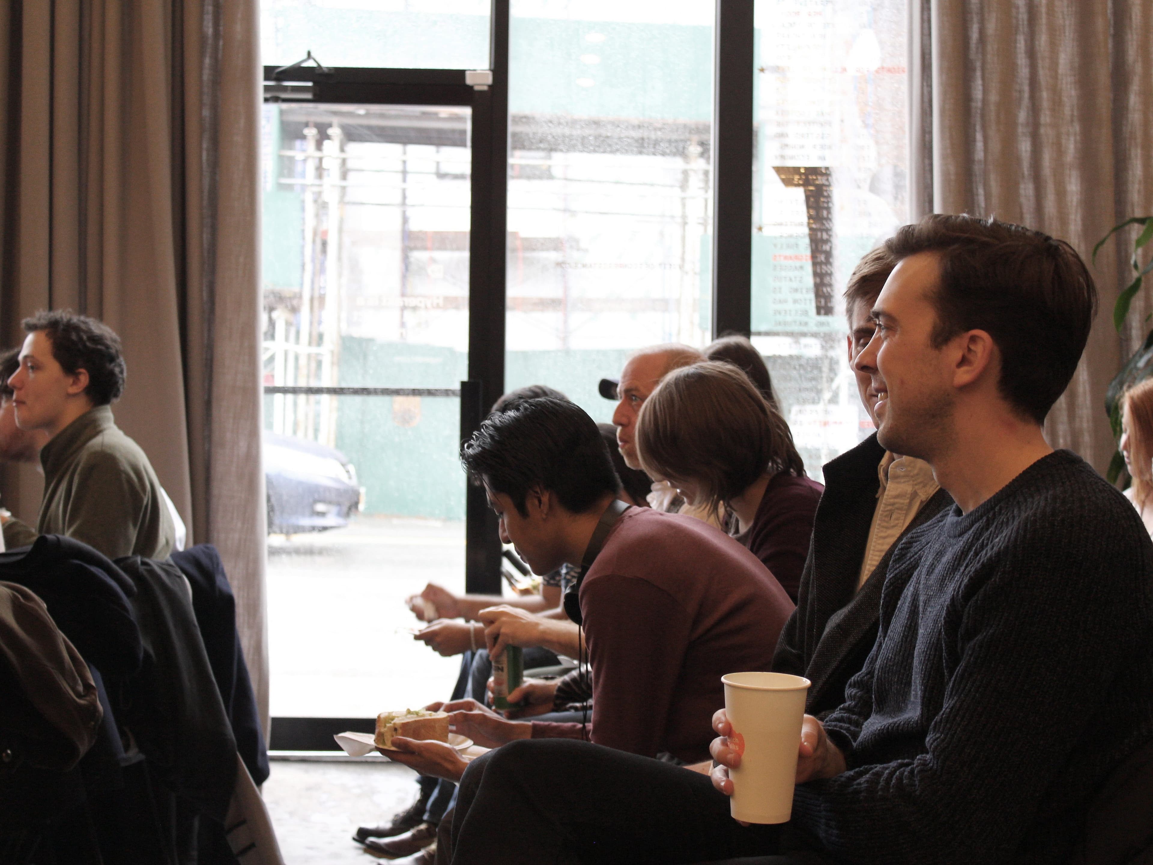 A group of people sitting in a room, attentively listening to a presentation. Some are holding disposable coffee cups and food. A man in the foreground is smiling. The room has large windows with sheer curtains, and a construction site is visible outside.