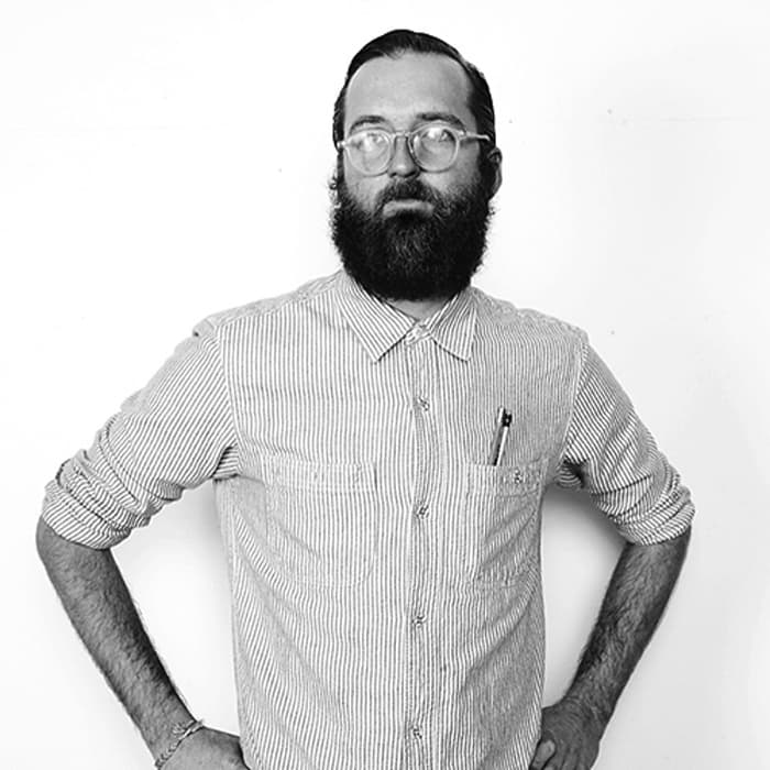 A bearded man wearing glasses stands against a plain white background. He has a serious expression, and his striped, button-up shirt has a pen in the chest pocket. He stands with his hands on his hips.