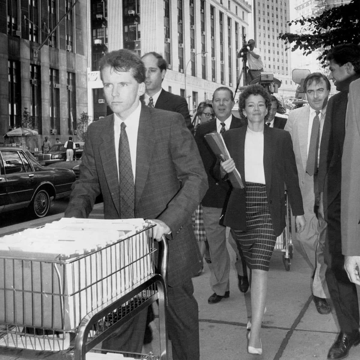 A group of people in business attire walk along a city street next to a building. One man pushes a cart filled with documents. The scene is bustling, with several pedestrians and a camera tripod visible in the background. The image is in black and white.