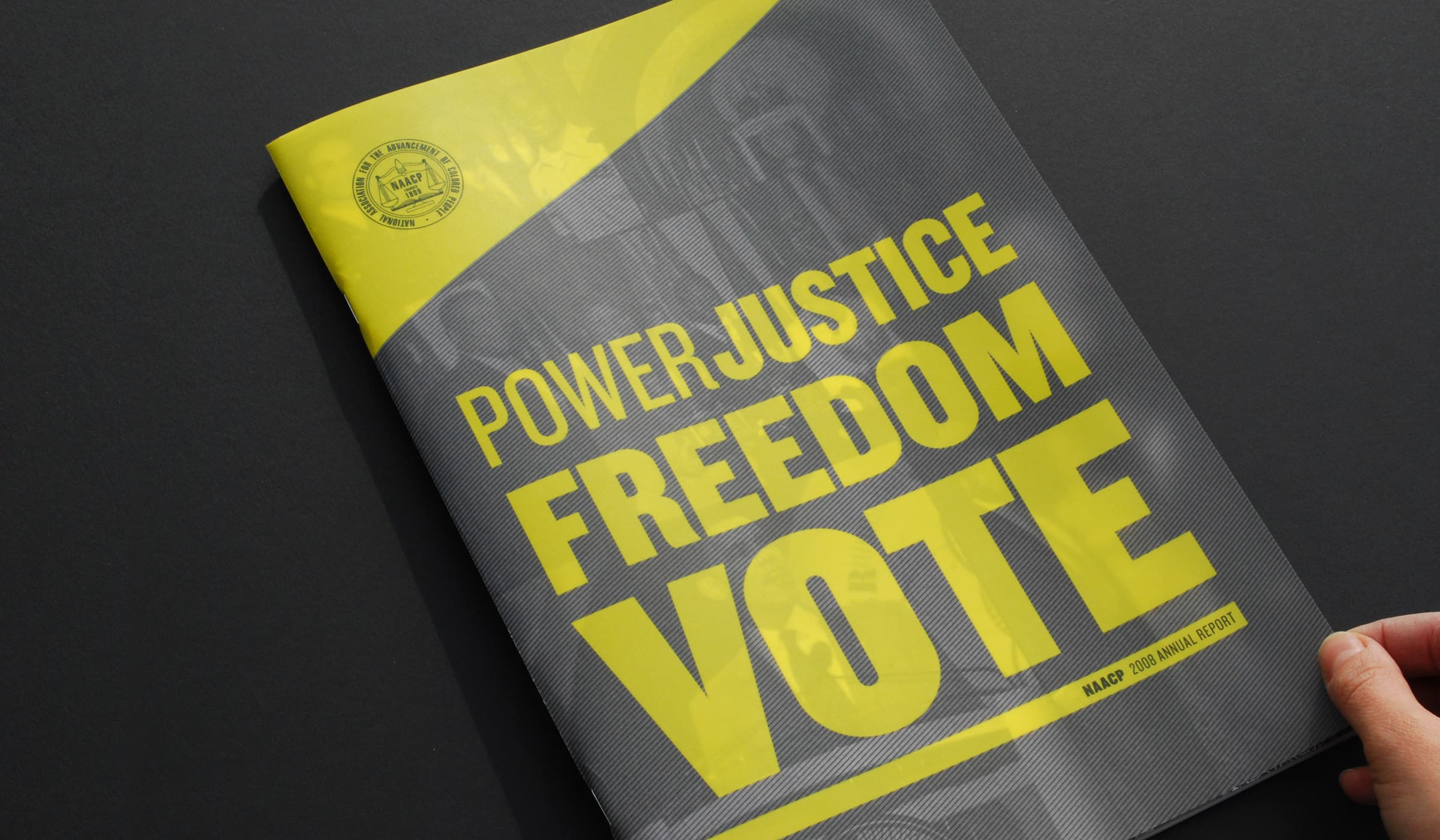 A hand holding a booklet with a yellow and gray cover. The cover features bold yellow text that reads "POWER JUSTICE FREEDOM VOTE." The top left corner shows an emblem, and the bottom right corner notes "NAACP 2014 Annual Report." The background is dark gray.