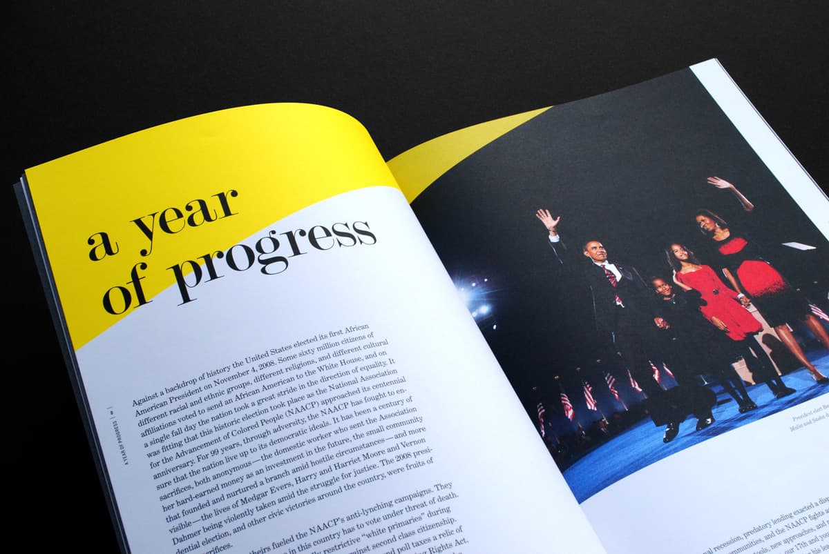 An open magazine shows a two-page spread. The left page, with a bright yellow top section, has black text stating "a year of progress" and an article below. The right page displays a photo of several people, including a man waving, against a dark background with American flags.
