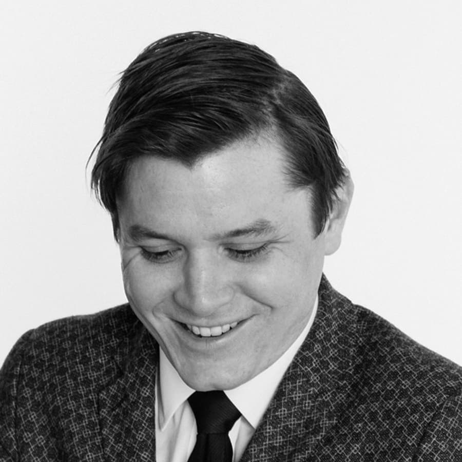 Black and white portrait of a smiling person with short, dark hair, dressed in a patterned jacket, white shirt, and dark tie. The person is looking downward with a slight smile, giving the image a candid and warm feel. The background is plain white.