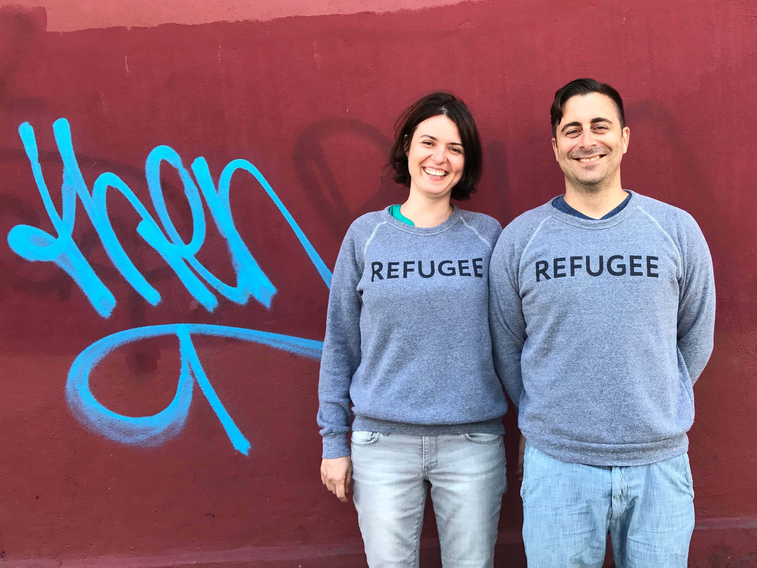 A smiling woman and man stand side by side against a red wall with blue graffiti. They both wear gray sweatshirts with the word "REFUGEE" printed on them in black text. The woman has short brown hair and the man has short dark hair.