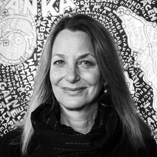 A black-and-white photo of a woman with long, straight hair smiling at the camera. She is wearing a dark top and standing in front of a wall covered with various words and patterns, providing a busy and artistic backdrop.