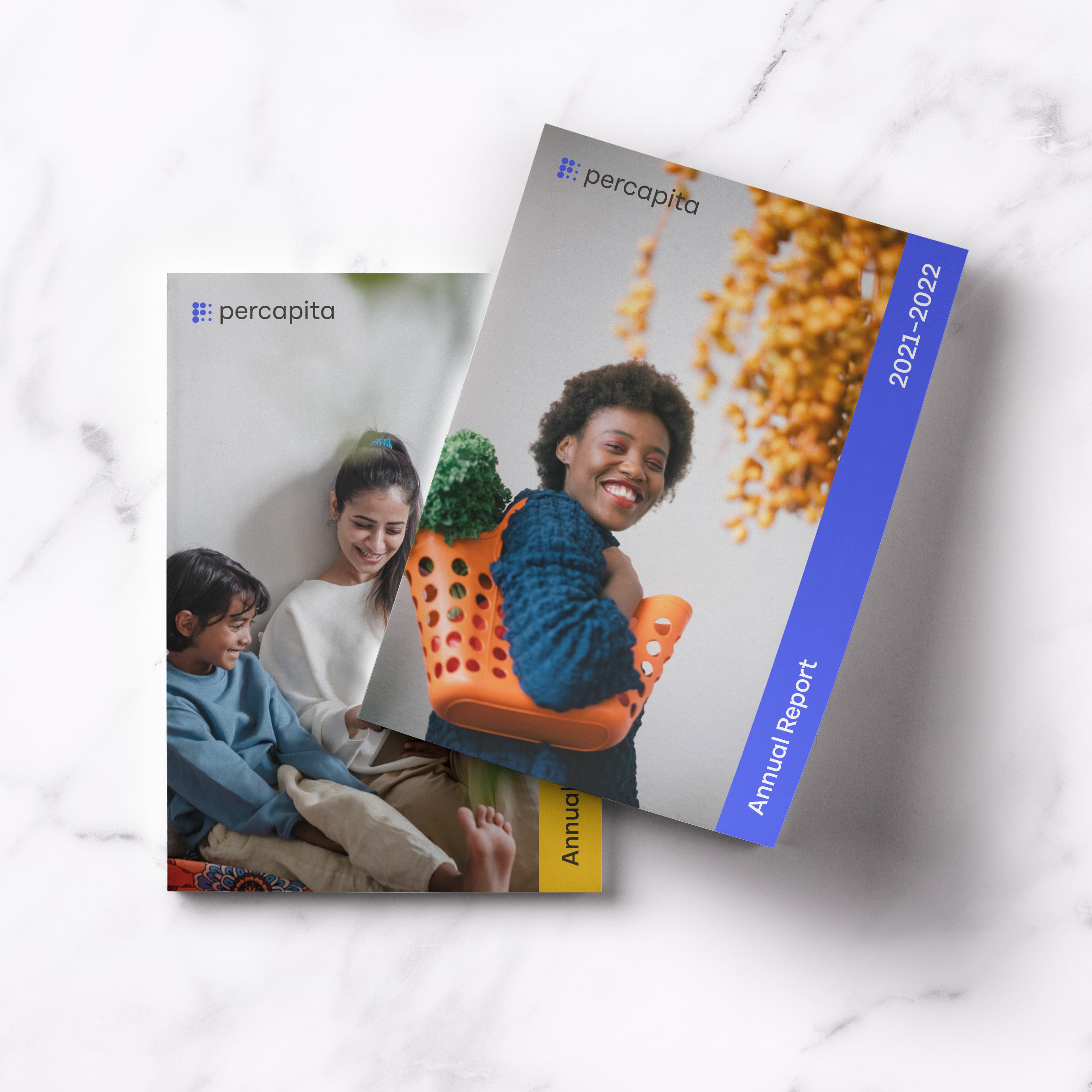 Two annual reports from "percapita" lie on a white marble surface. The top report, for 2021-2022, features a smiling person in a knitted sweater with colorful pom-poms. The bottom report shows a person reading with a child, both looking content.