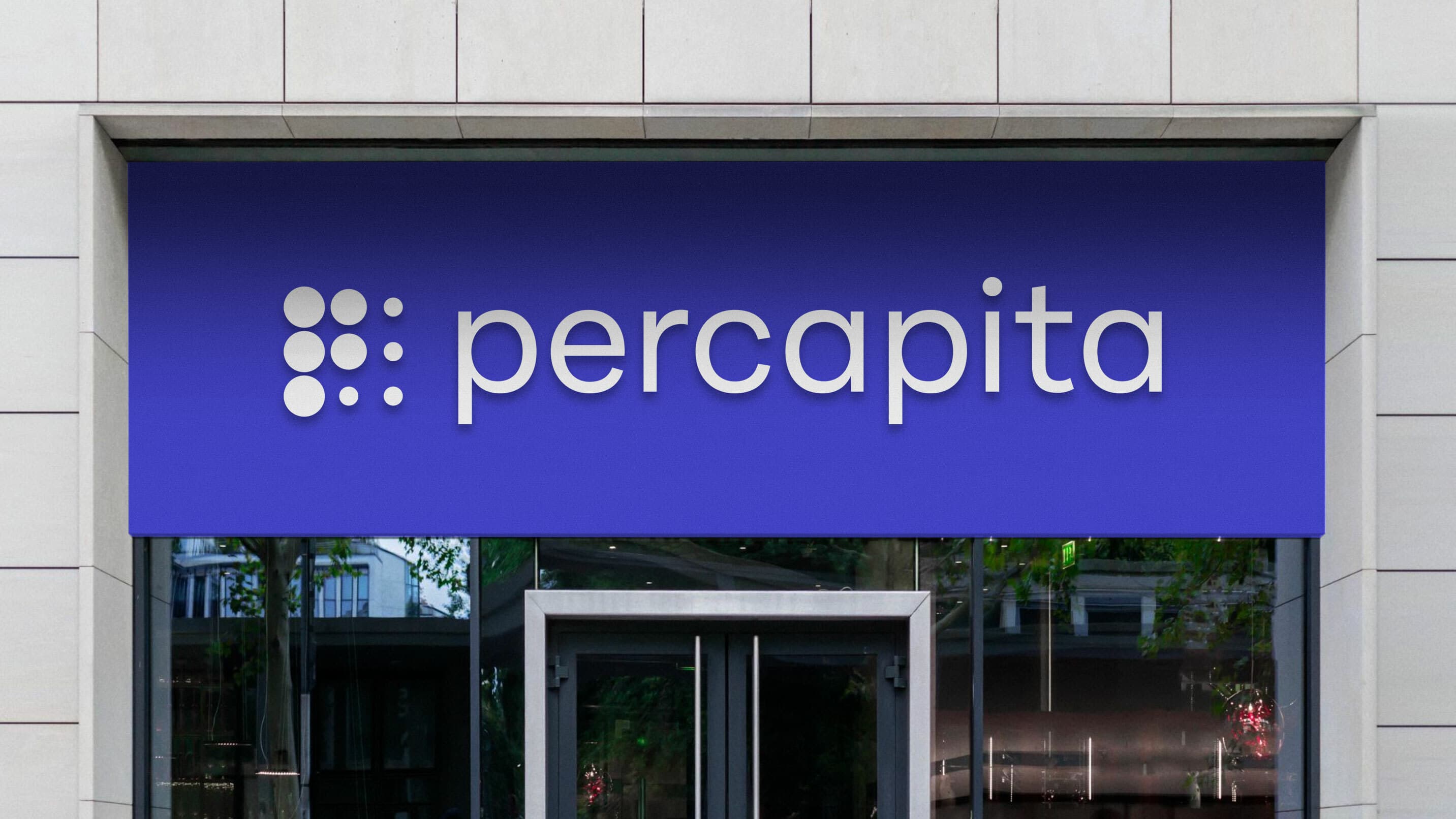 A storefront with a blue sign above the door. The sign displays a logo consisting of circular dots arranged in a grid pattern next to the text "percapita" in modern, lowercase white letters. The building exterior is light-colored with large glass windows.