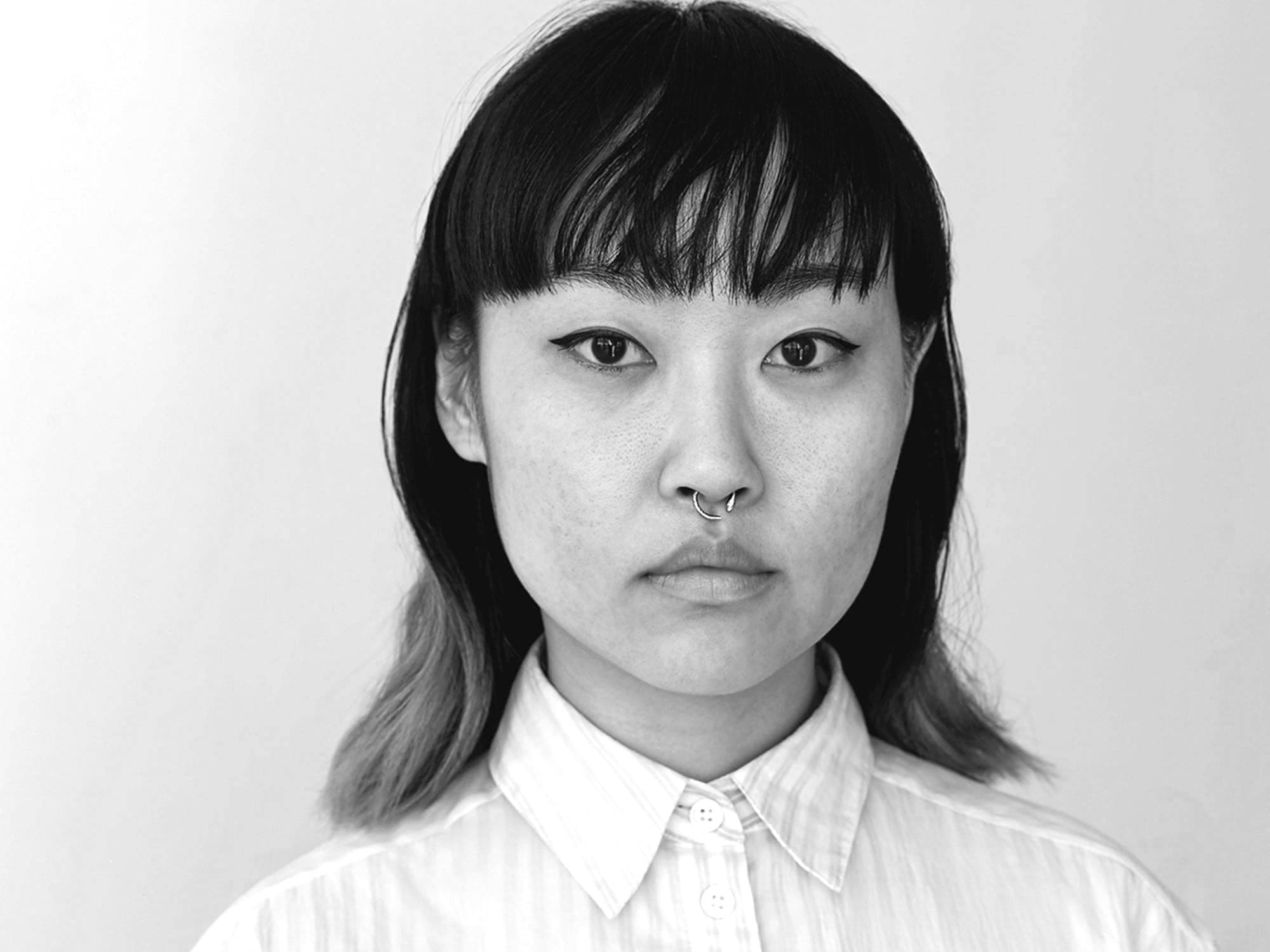 Black and white portrait of a person with shoulder-length hair that is darker on top and lighter at the ends. They have straight-across bangs, are wearing a collared shirt, and have a septum piercing. They are looking directly at the camera with a neutral expression.