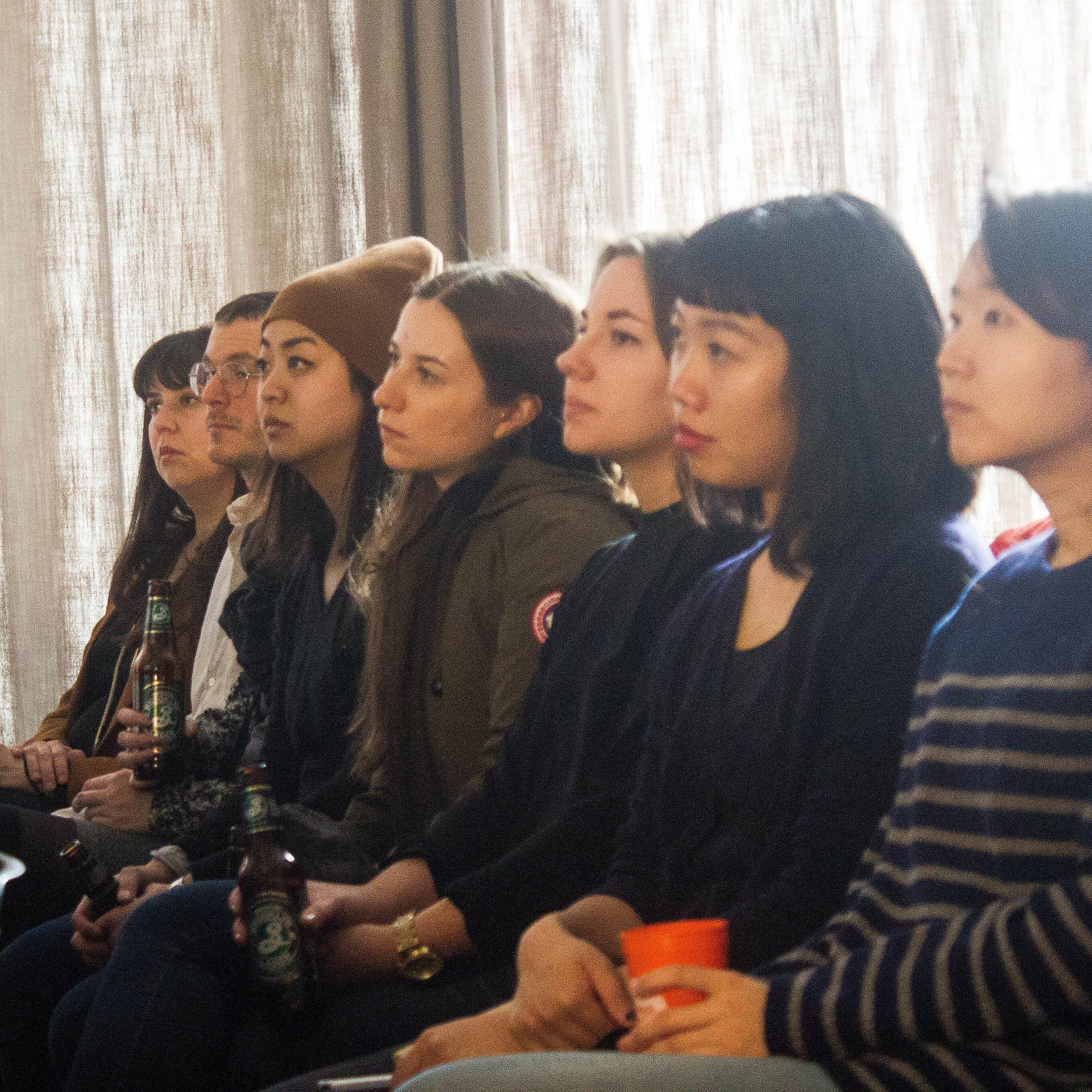 A group of people sitting in a row, attentively listening to a speaker. The setting appears to be a lecture or seminar room with beige curtains in the background. Some individuals are holding drinks. The atmosphere is focused and engaged.
