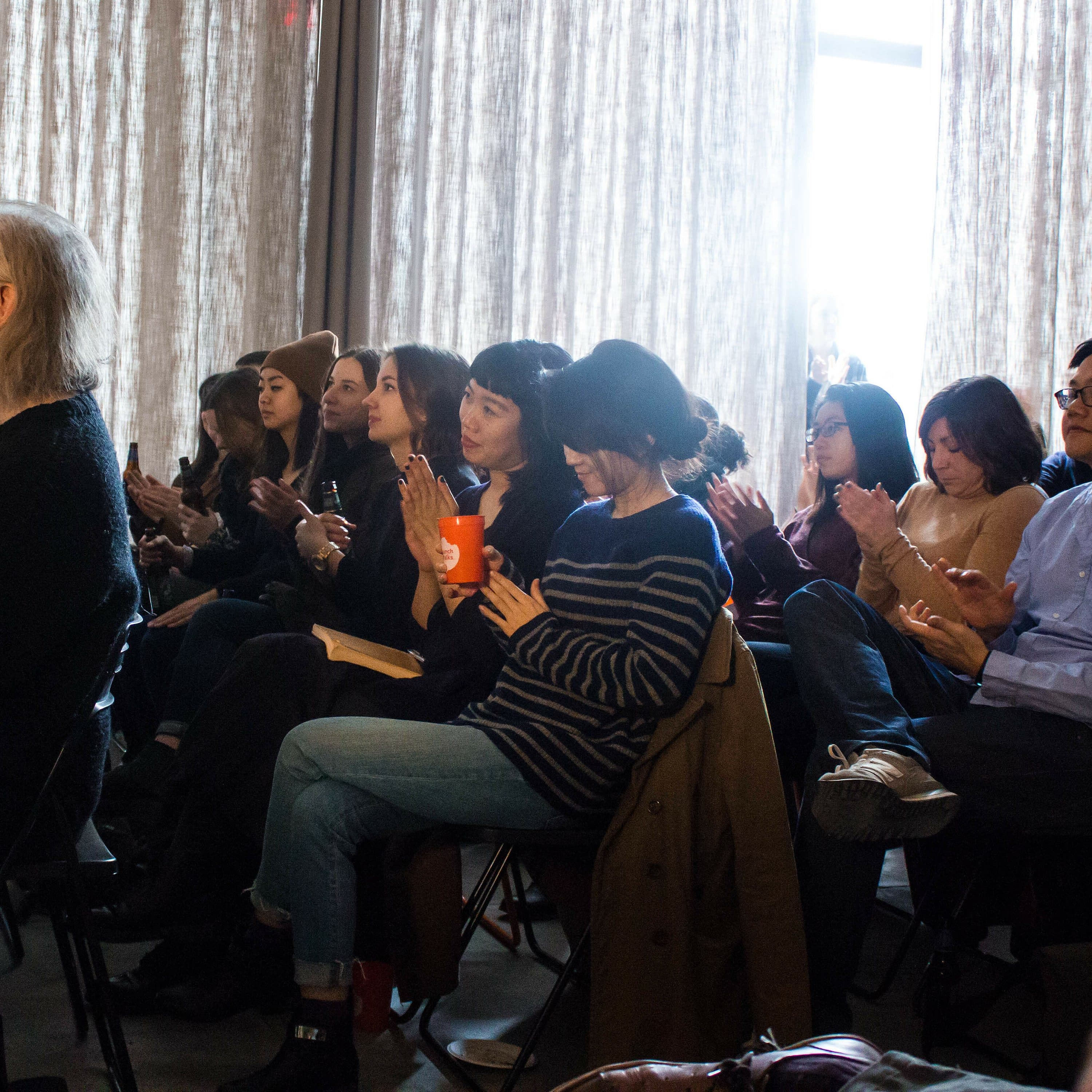 A diverse group of people seated in rows are attentively listening and clapping in a room with light-colored curtains. The audience includes individuals of various ages and backgrounds. The room is softly illuminated with natural light coming through the windows.