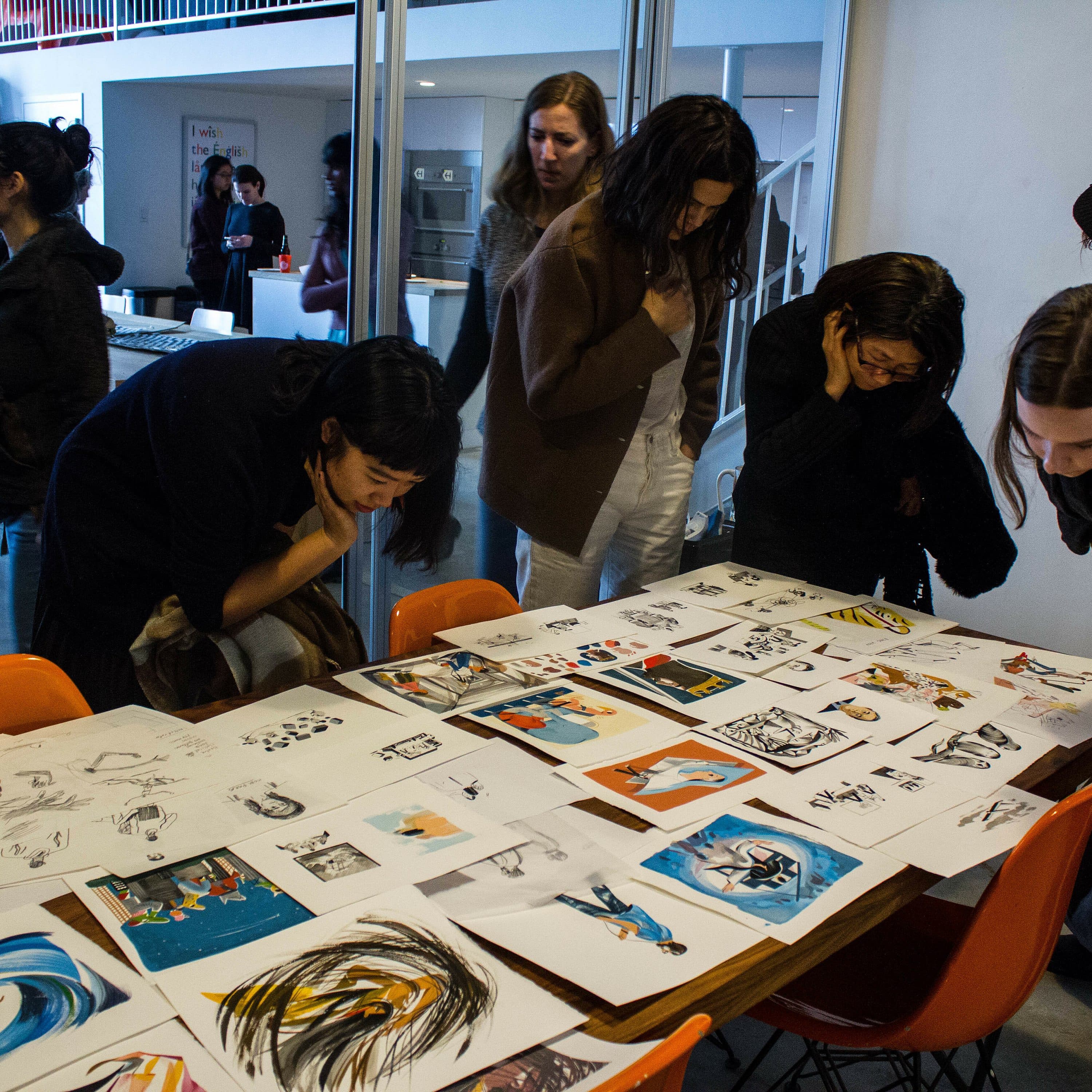 A group of people is gathered around a table covered with various artworks in a gallery-like setting. They are intently observing the drawings and paintings, which feature a range of subjects and styles. Others in the background are engaged in conversation.