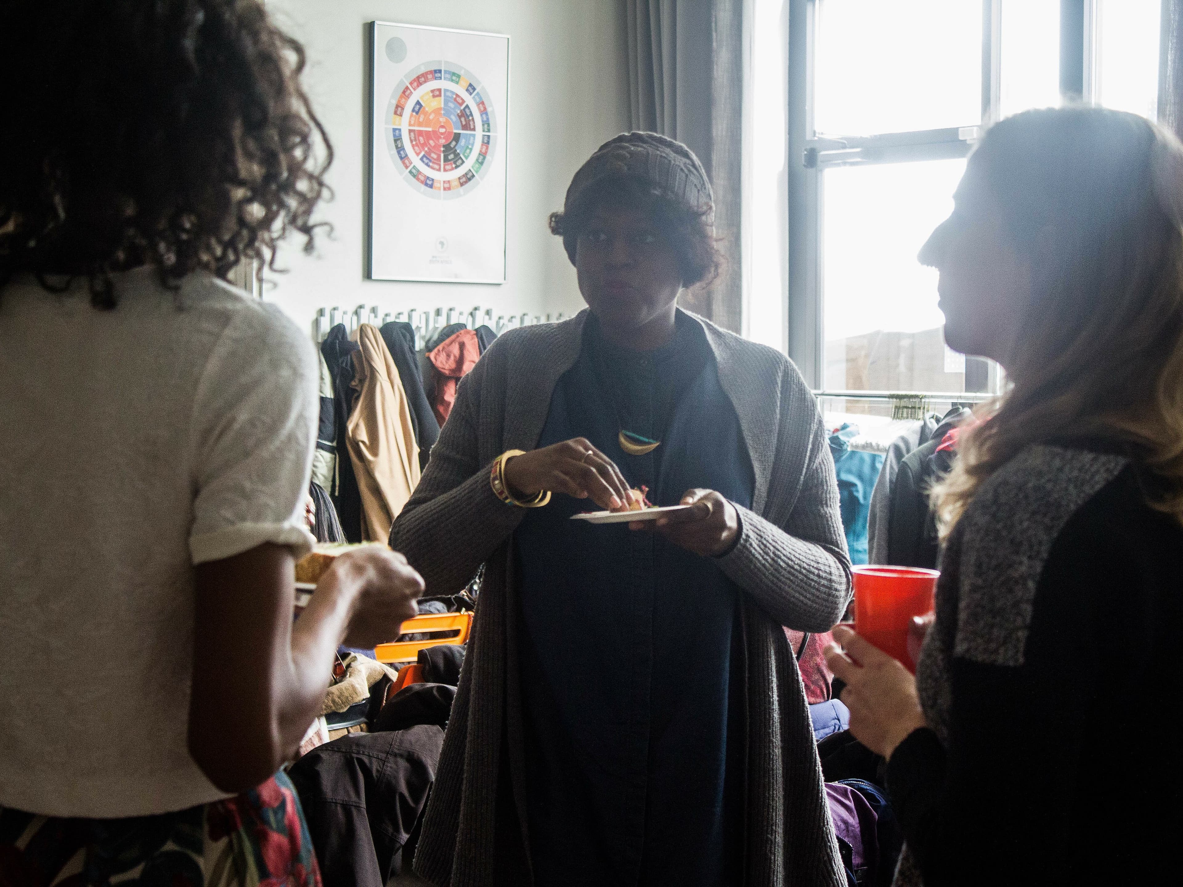 Three people are engaged in conversation at a casual indoor gathering. One person in the center is holding a plate of food, while the other two listen. Coats and jackets hang on hooks in the background, and a window allows natural light into the room.