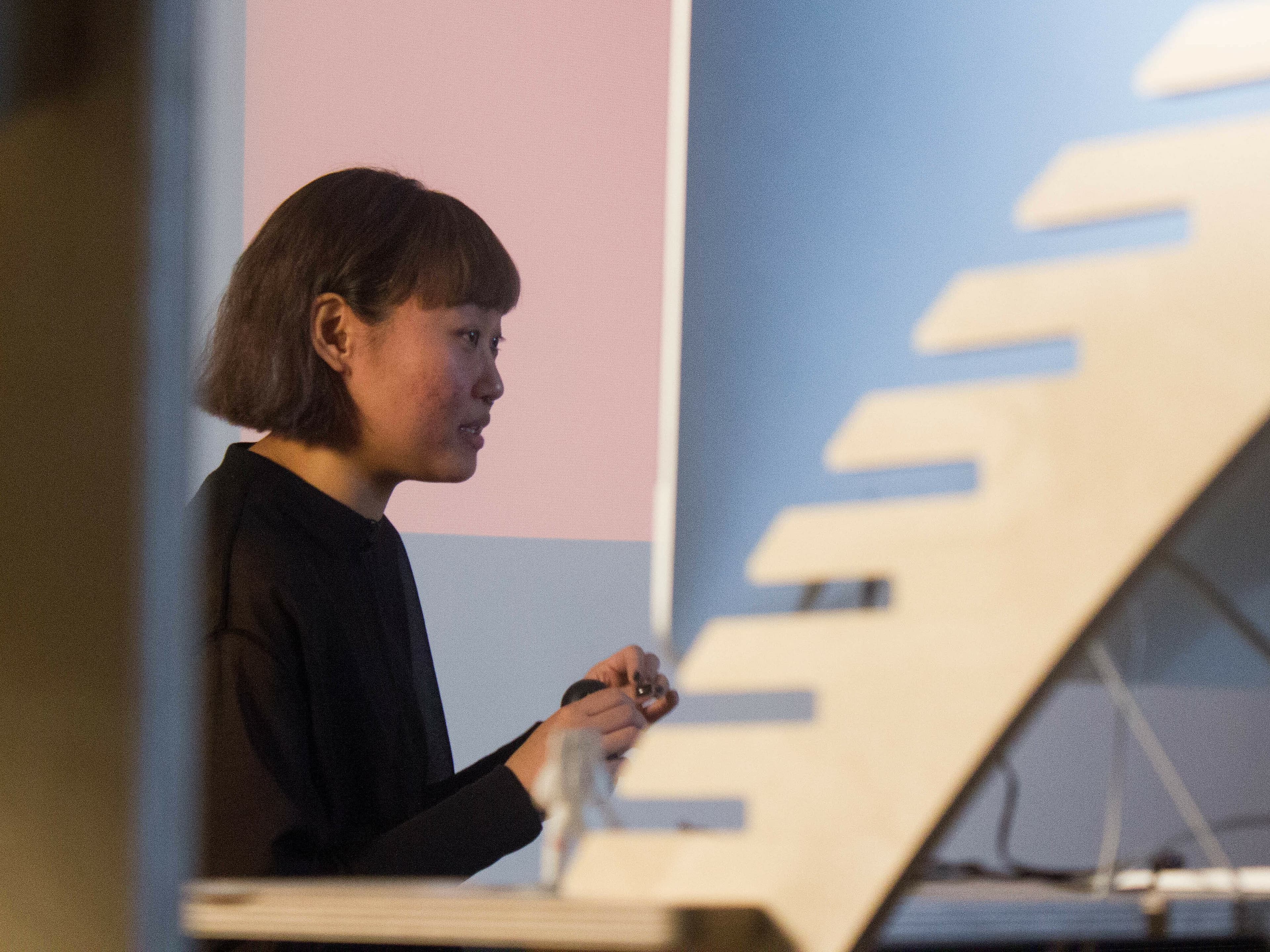 A person with short hair is standing and holding an object in their hands, facing towards the left. They are indoors, and part of a stairway is visible in the foreground. The person appears to be engaged or focused on something outside the frame.