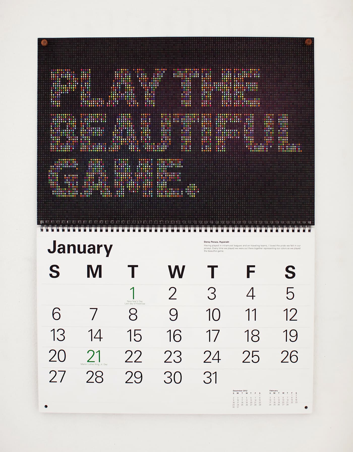 A calendar hanging on a white wall. The top half features colorful text reading "PLAY THE BEAUTIFUL GAME." The bottom half displays the month of January with dates and days of the week, starting with Sunday. Several dates, including 1 and 21, are highlighted in green.