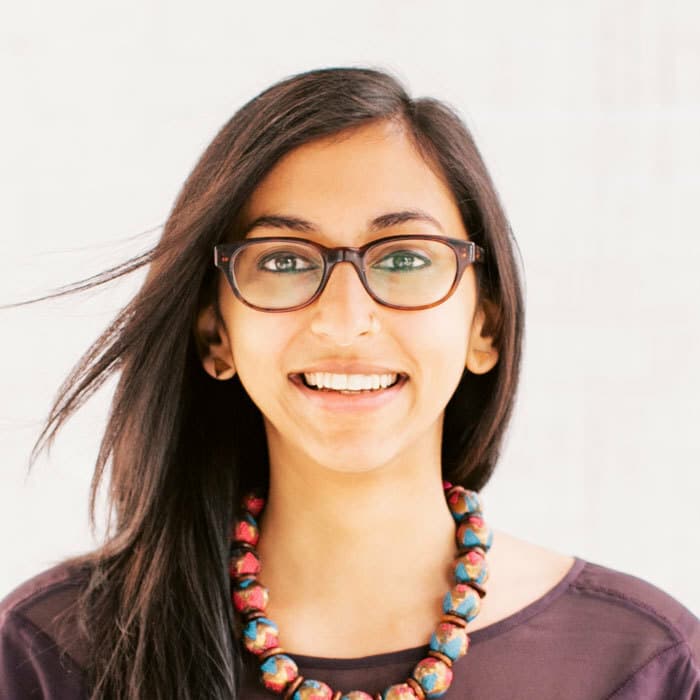 A woman with long dark hair and glasses is smiling, facing the camera. She is wearing a dark top and a colorful beaded necklace. The background is plain and light-colored.