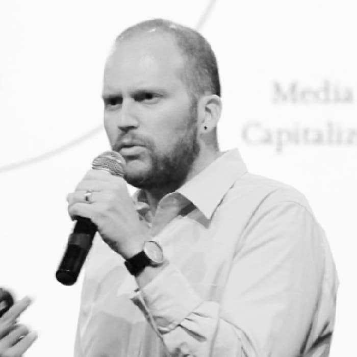 Black and white image of a person with a beard and short hair, wearing a light-colored shirt, speaking into a microphone. In the background, there is a blurred out screen or projection with some text. The person appears to be making a gesture with their other hand.