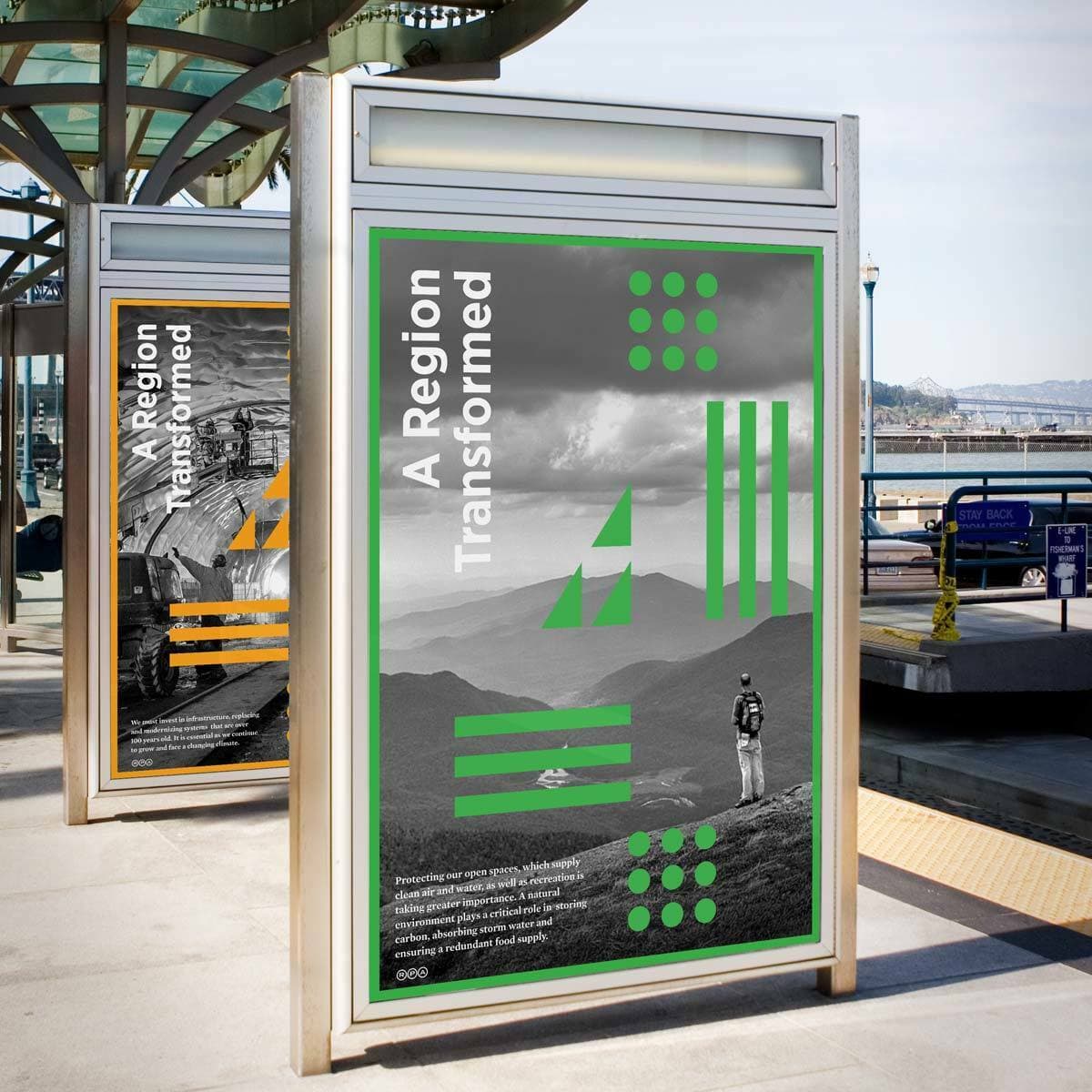 Outdoor posters at a bus stop. The prominent poster has the title "A Region Transformed" and displays a person hiking in a mountainous area with geometric designs overlay. Another similar poster is visible in the background. A harbor view is in the distance.