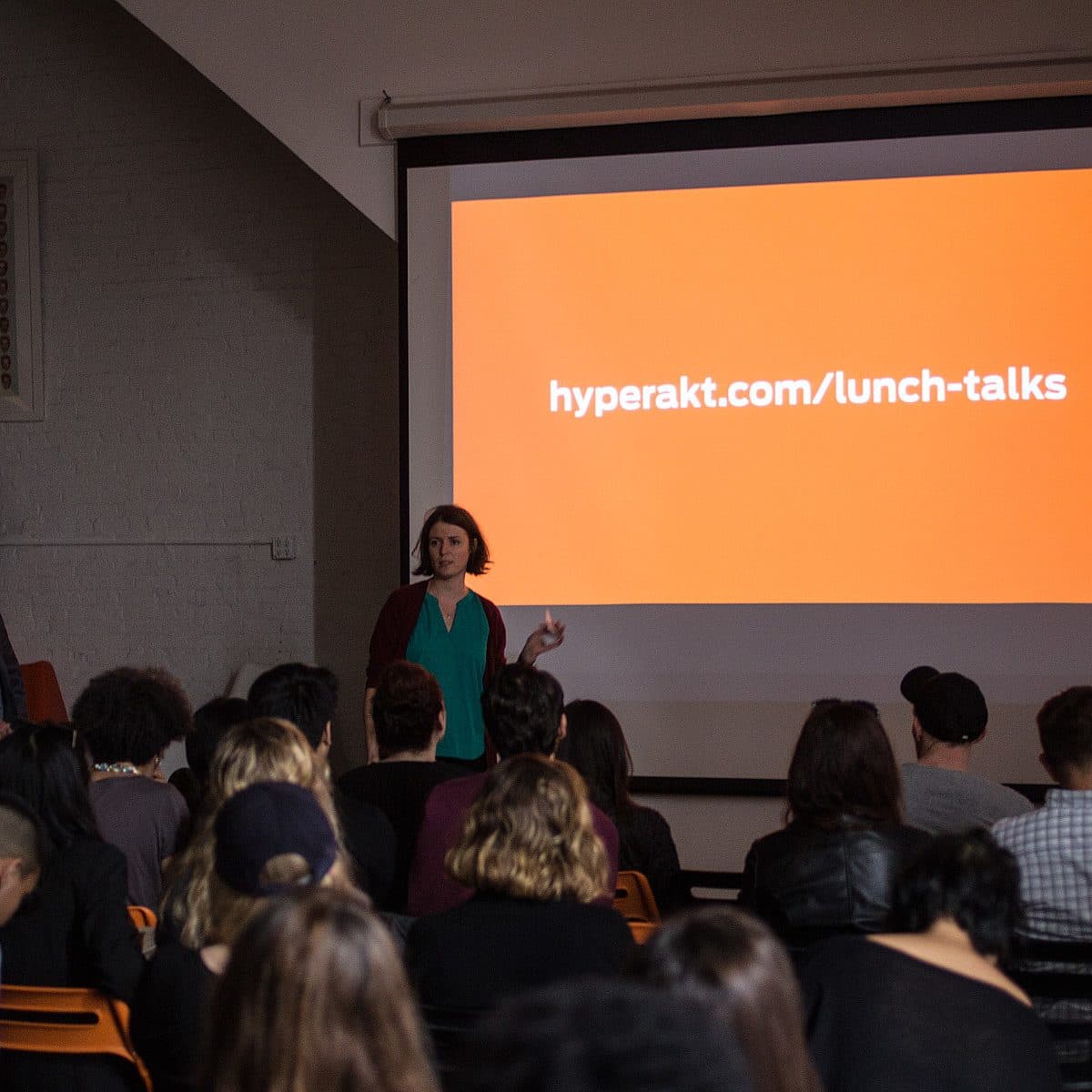 A woman stands in front of an audience, gesturing while speaking. A man stands nearby watching. Behind them is a large screen displaying the text "hyperakt.com/lunch-talks" on an orange background. The audience is seated and attentive.
