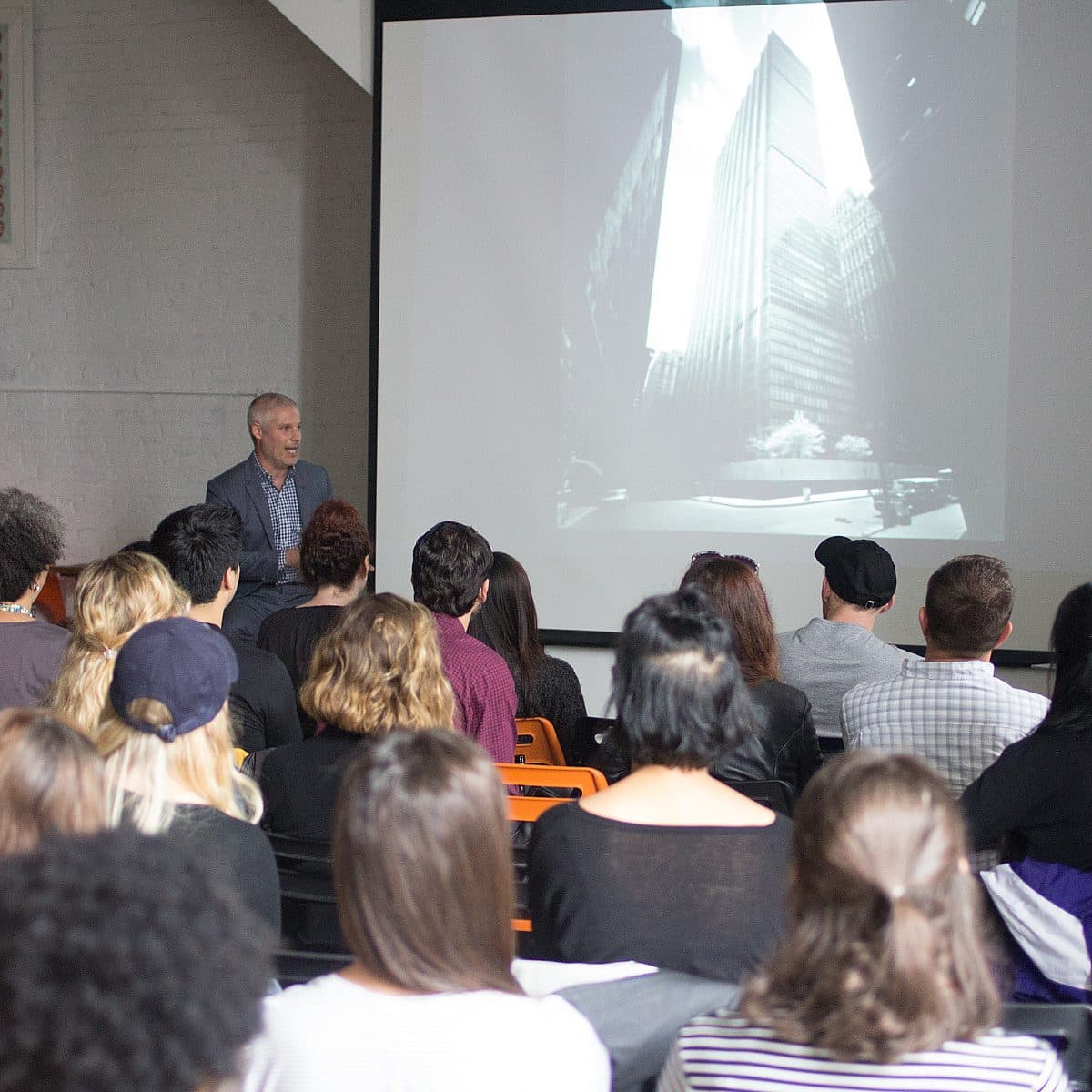 A group of people sits facing a large screen displaying a black-and-white photo of tall buildings. A speaker stands to the left of the screen, presenting to the audience. The room has a modern interior, with a computer setup visible in the background.