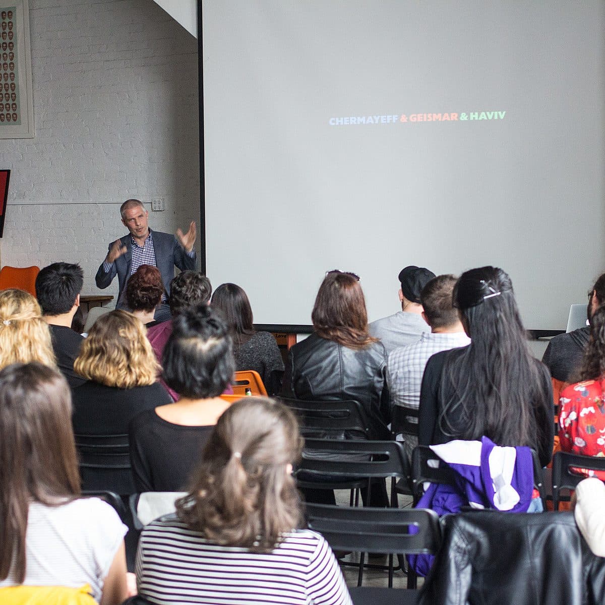 A group of people sit facing a speaker in a room, watching a presentation on a large screen. The speaker is gesturing with both hands. The screen displays the words "CHERMAYEFF & GEISMAR & HAVIV" in different colors. The audience is attentive, with some taking notes.