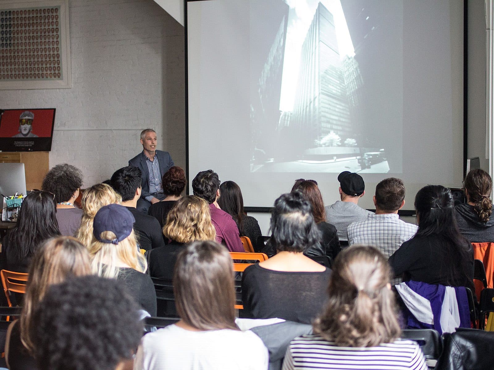 A speaker stands at the front of a room, presenting to an audience. The presentation screen behind him displays a black-and-white photo of tall buildings. The audience members, seated in rows, are listening attentively. A laptop and some equipment are visible on a table.