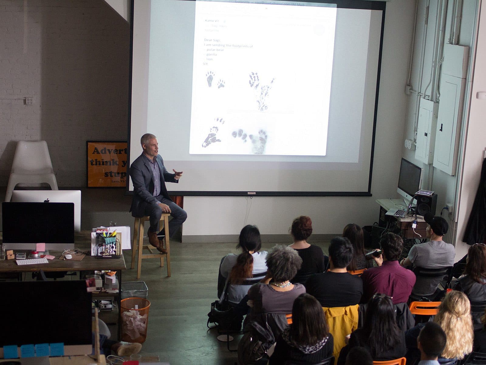 A man is giving a presentation to an audience in a modern office space. He is sitting on a stool in front of a large projector screen displaying text and images. The audience is seated and attentively listening. Office equipment and supplies are visible in the foreground.