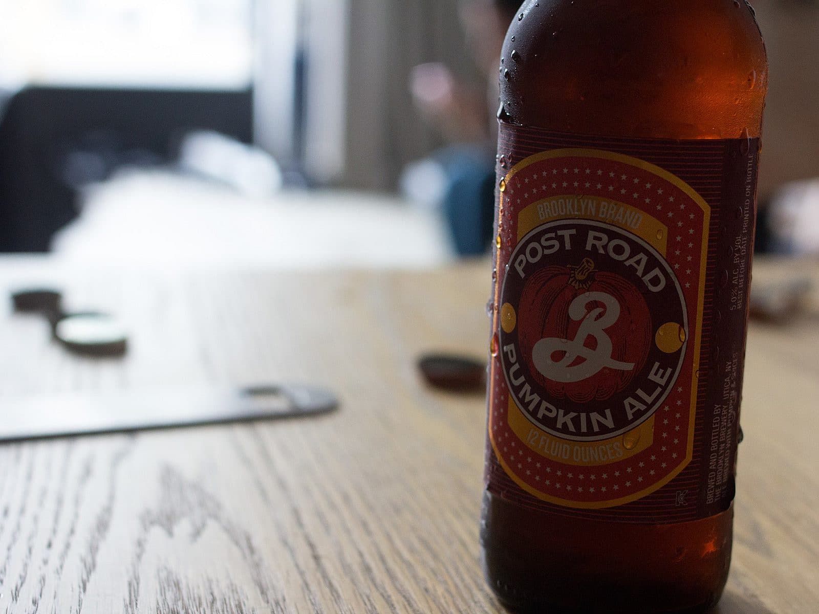 A bottle of Brooklyn Brand Post Road Pumpkin Ale sits on a wooden table. The label is red and yellow with white text and a pumpkin illustration. In the background, the image is out of focus, showing some coins and a blurred figure sitting by a window.