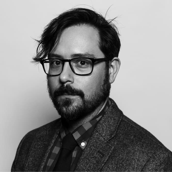A black and white portrait of a man with glasses, a beard, and mustache. He has short, wavy hair parted to the side and is wearing a tweed jacket, checkered shirt, and dark tie. The background is plain and light-colored.