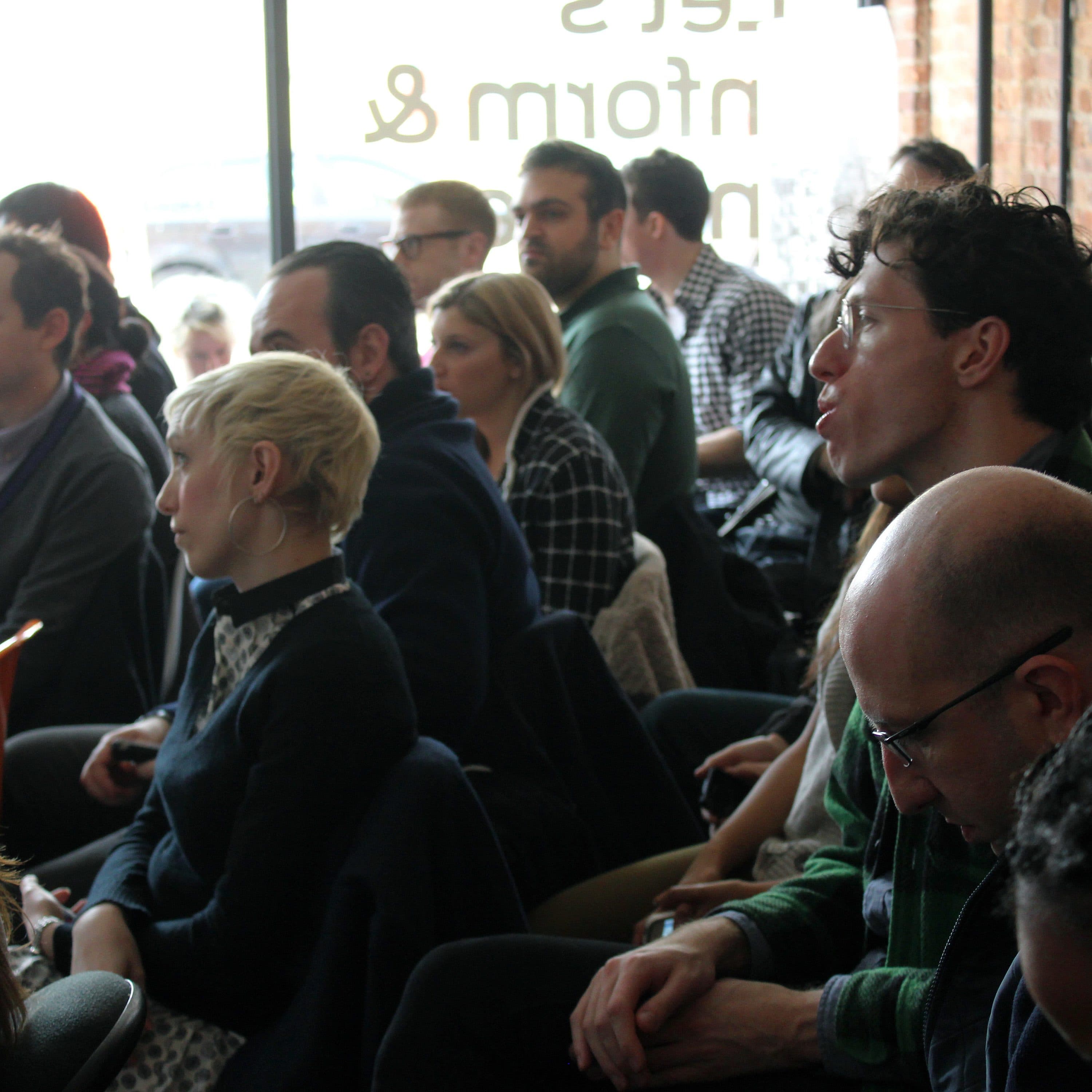 A group of people attentively listening in a seated arrangement within a well-lit room. The background features large windows and exposed brick walls. Some attendees are taking notes while others are engaged with the speaker off-camera.
